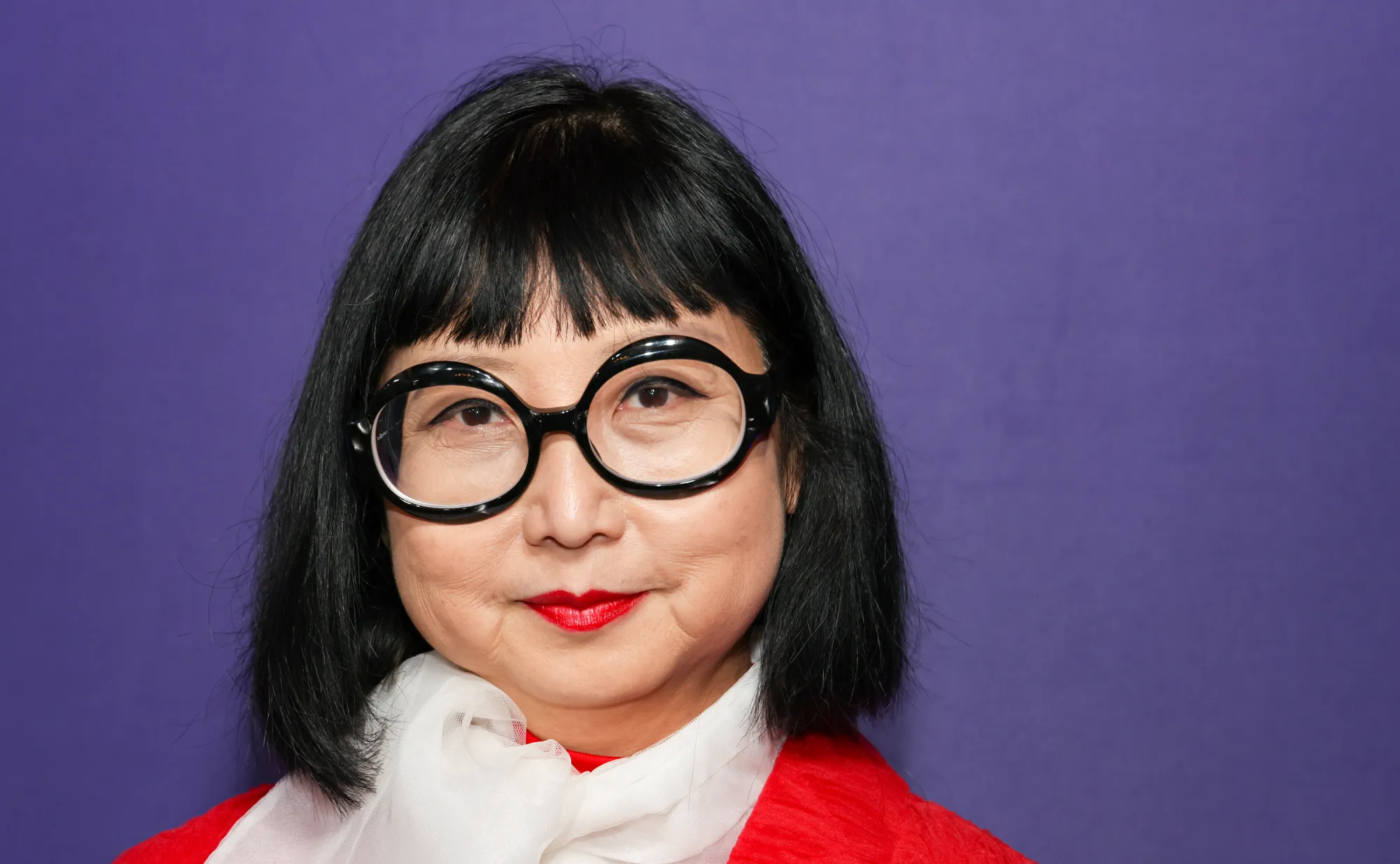 Costume Designer Shirley Kurata. She is wearing large, round black glasses, a white chiffon scarf and a red blouse that perfectly matches her red lipstick