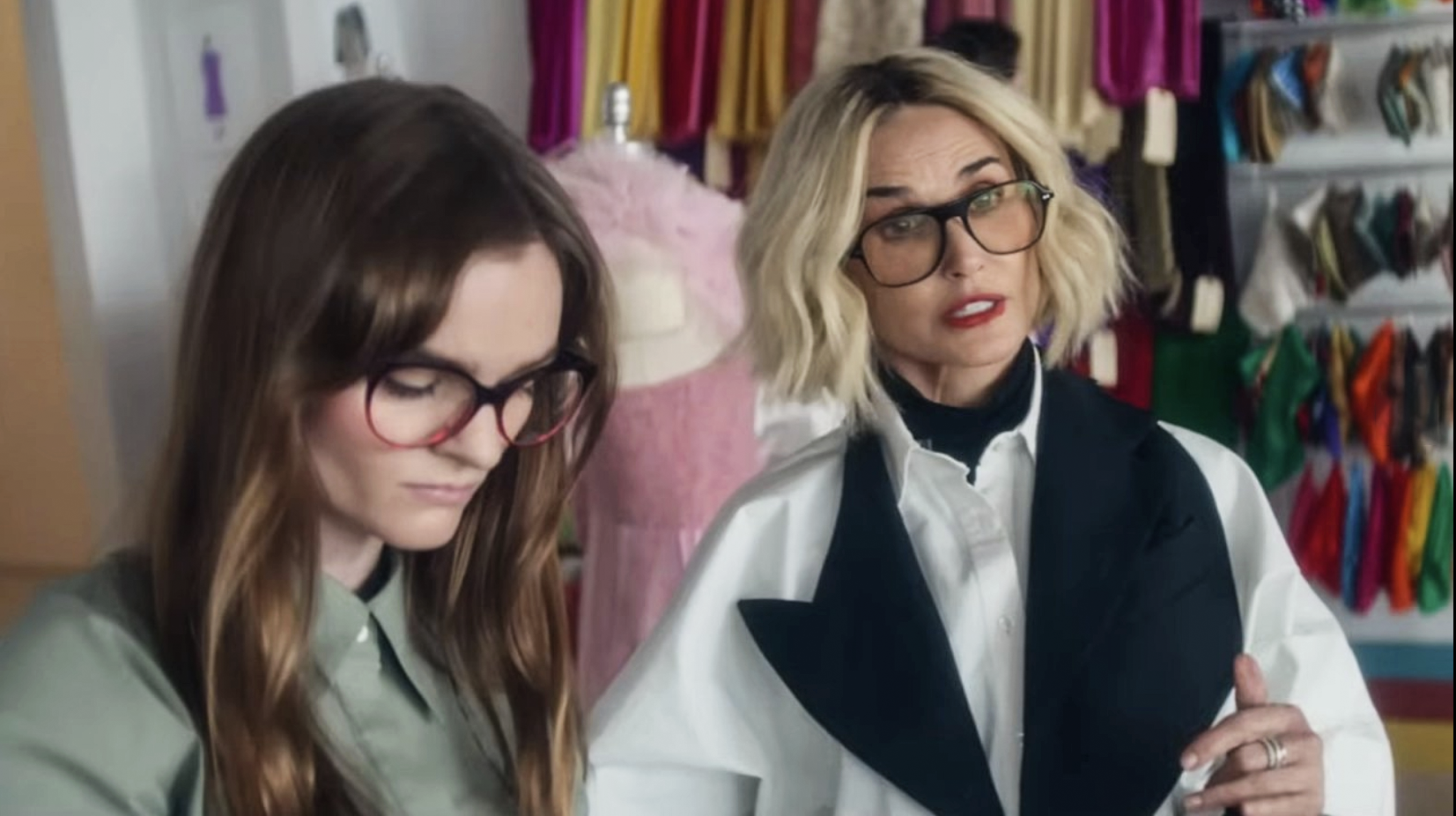 Kerris Dorsey on the left and Demi Moore on the right. Kerris Dorsey is wearing large round tortoise shell glasses. A khaki green blouse and is looking down at something. Demi Moore has a blonde bob with black roots showing, black large rimmed designer glasses, a white blouse with a white coat that has a huge lobbed black collar and a black cravat.