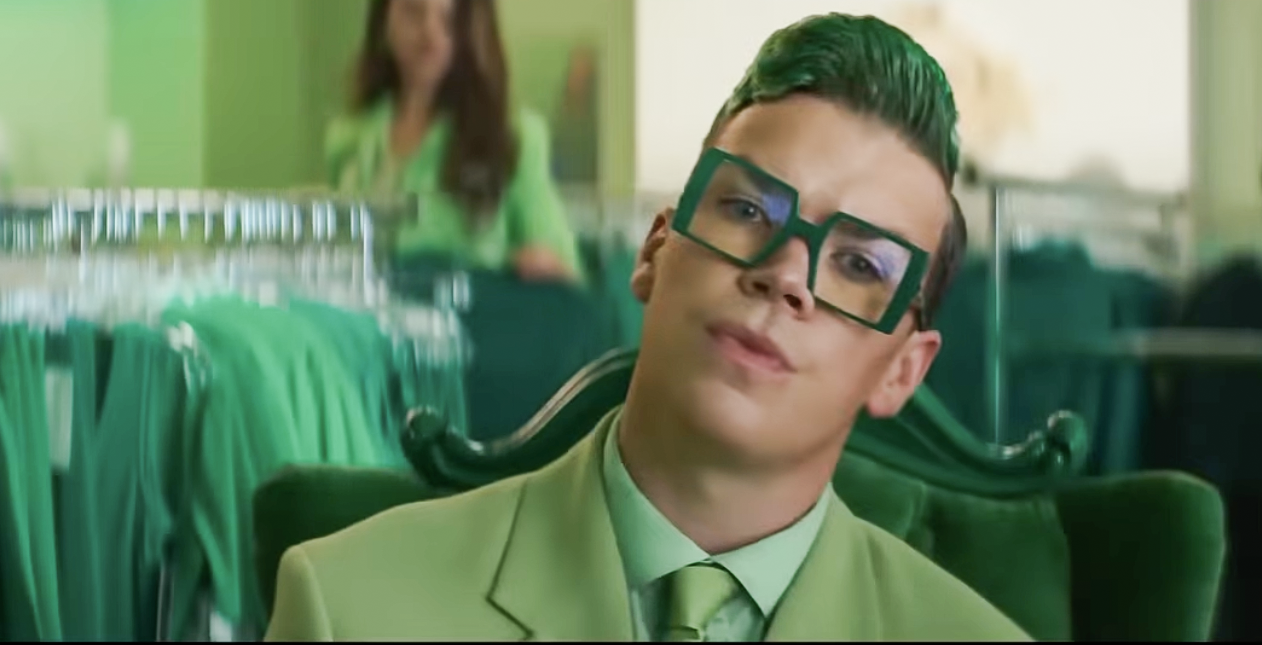 Will Poulter wearing boxy large glasses, and dressed in a spring green suite with a mint green shirt and spring green tie. With a green with a rack of clothes behind him.