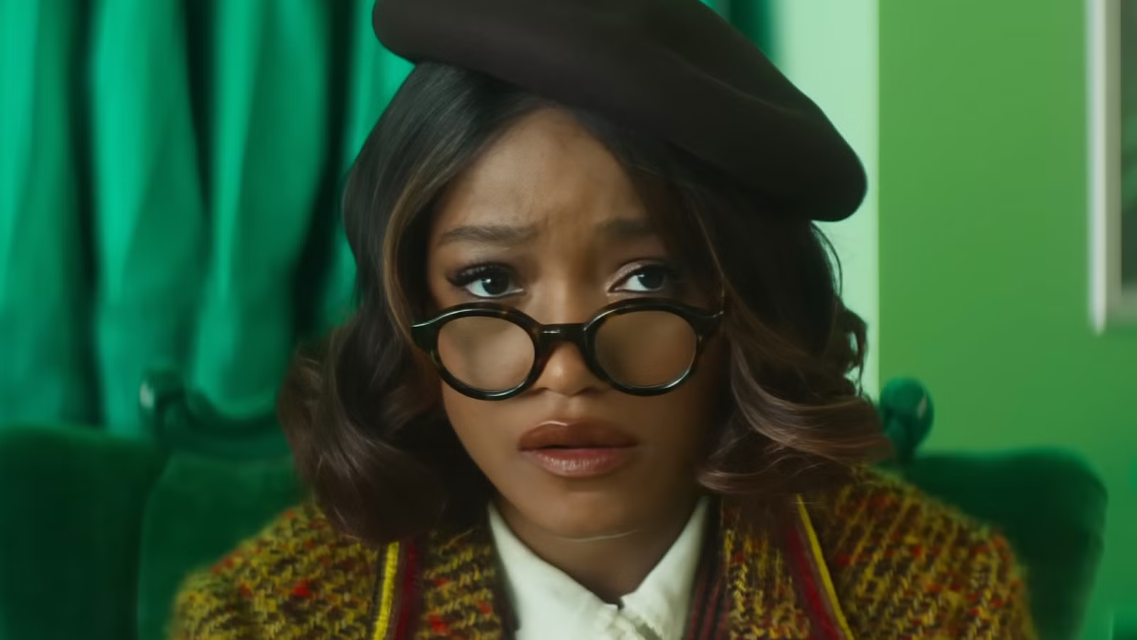 Keke Palmer with green background. Wearing stylish tortoise shell glasses, beret, white blouse under a brown wool jacket.