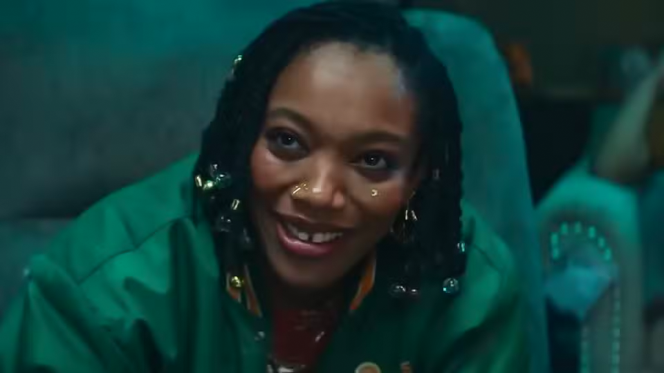 Naomi Ackie in Green jacket with green sofa and chairs behind her.