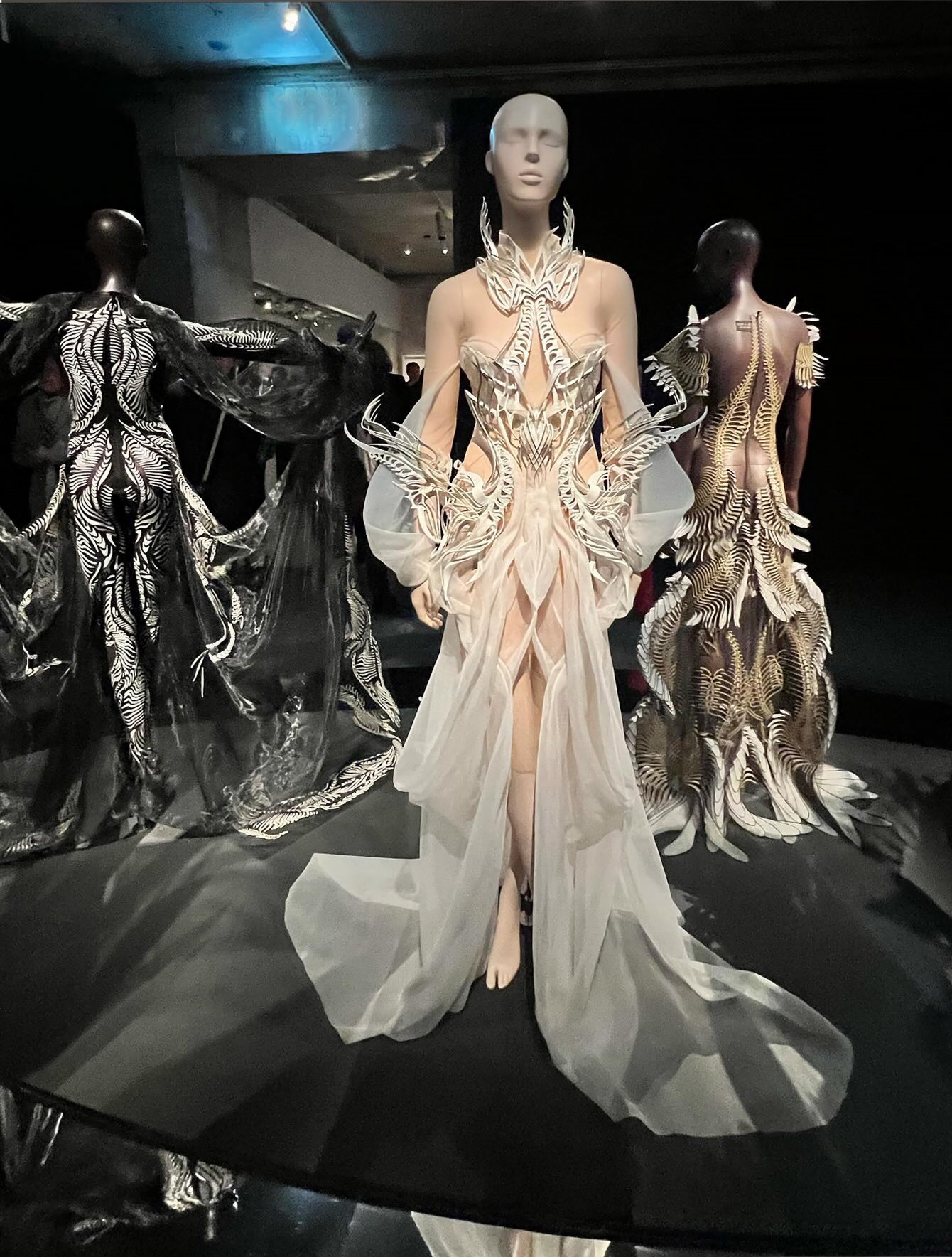 A museum gallery view with a mannequin wearing a white translucent Iris van Herpen gown with rib-like sculptural structures, surrounded by other black, white, and gold garments.