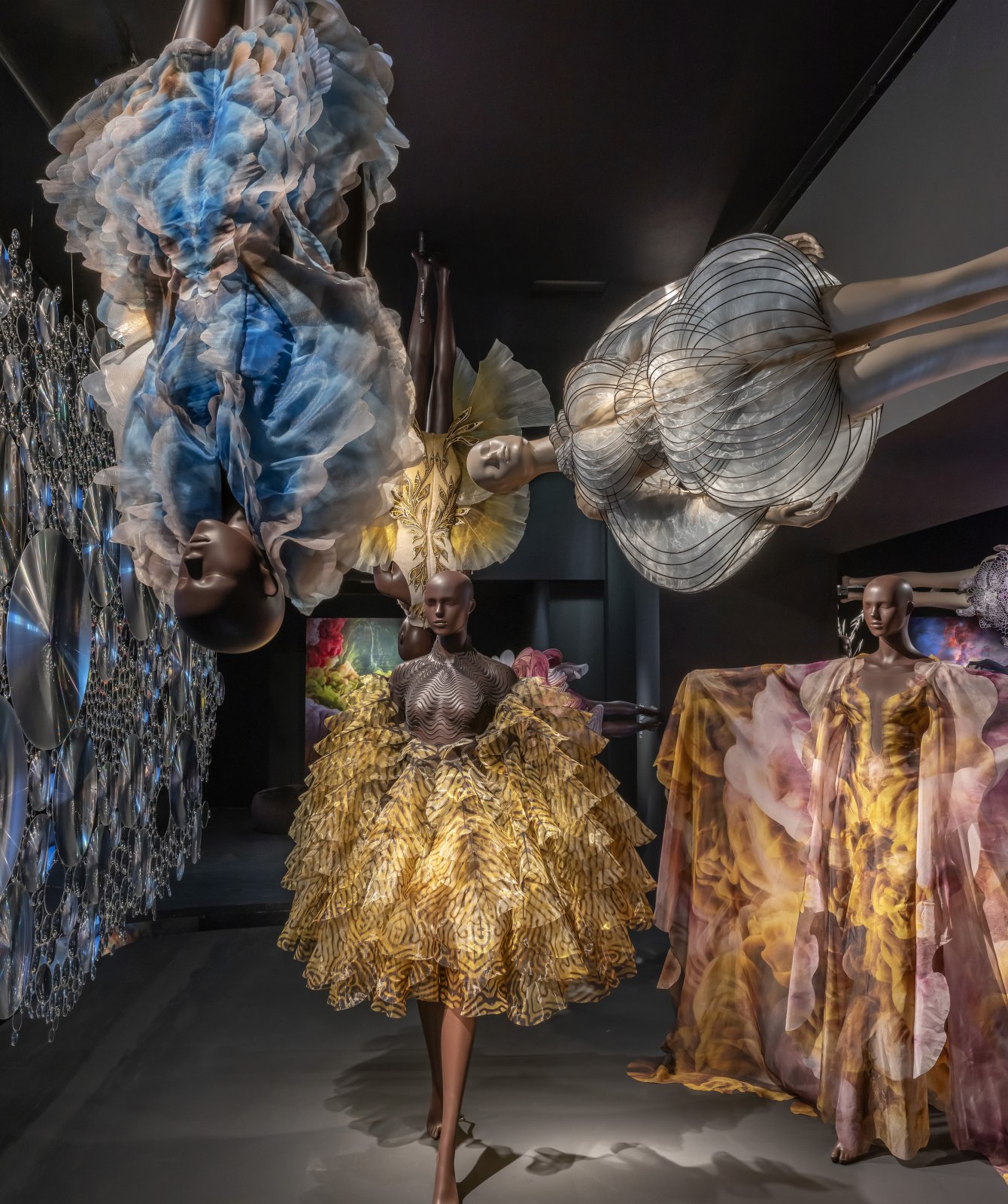 A museum installation view with multiple Iris van Herpen garments displayed on mannequins, including colorful translucent dresses and sculptural forms suspended and arranged throughout a dark gallery.