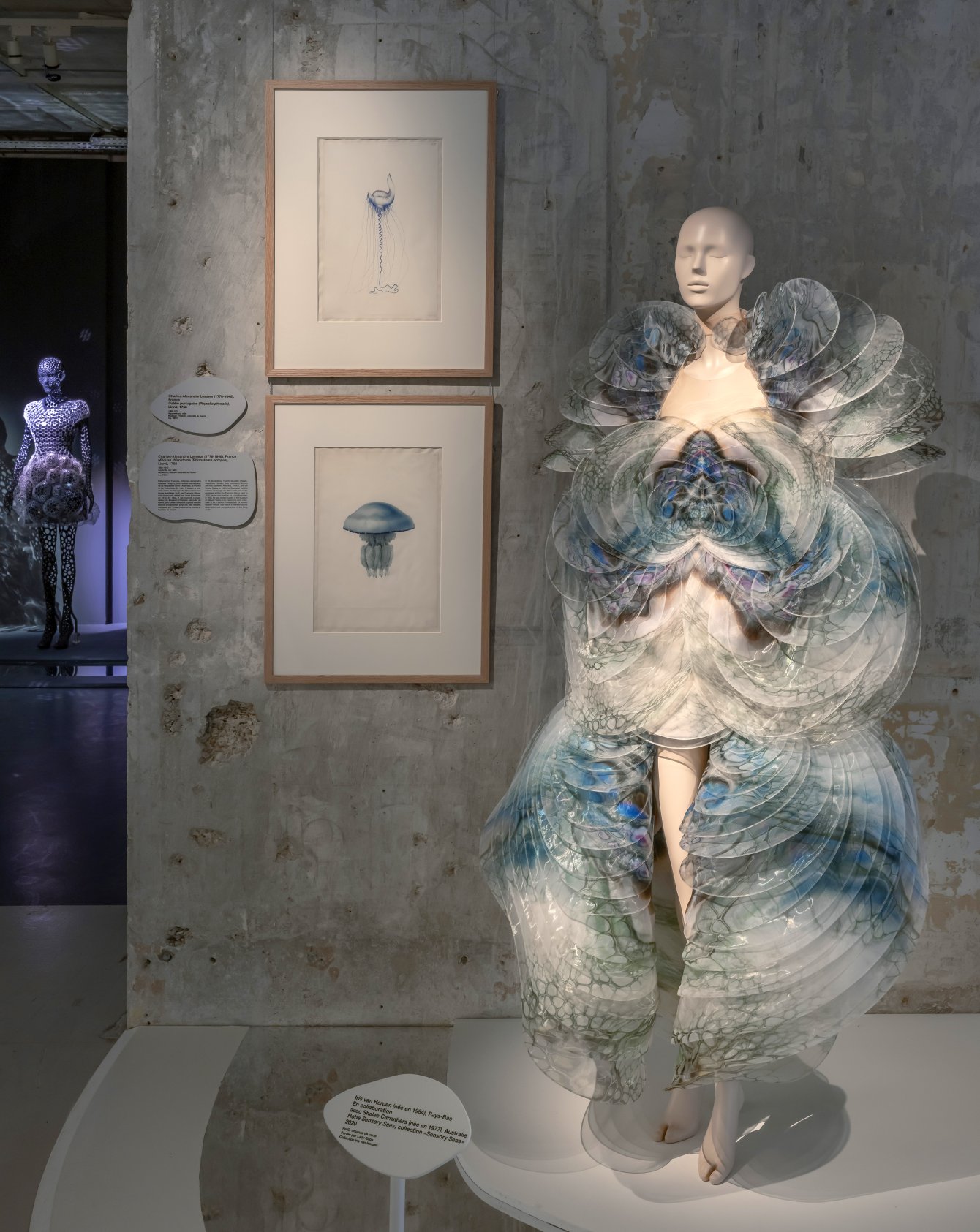 A dark gallery scene showing a mannequin in a black tubular Iris van Herpen garment displayed beside a classical bronze sculpture of a male figure.