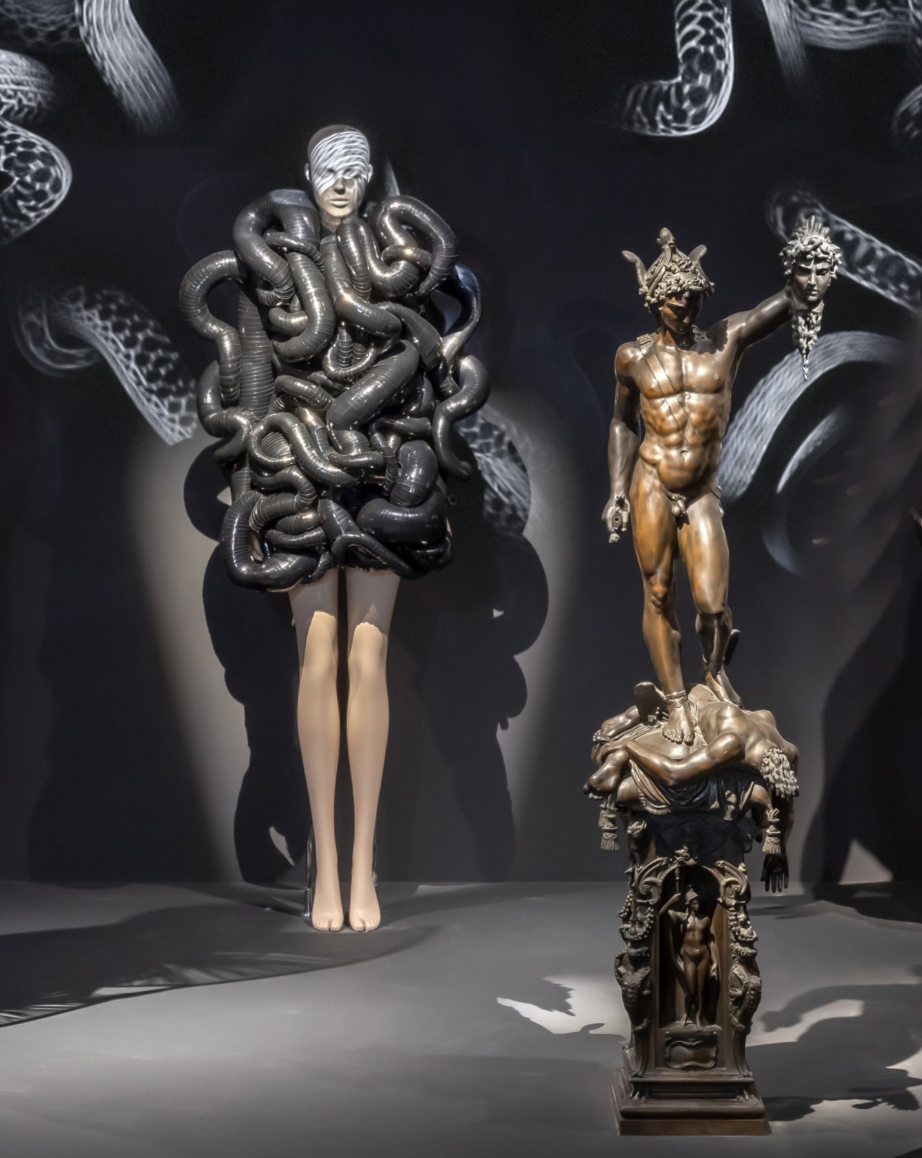 A museum installation with two mannequins in white sculptural Iris van Herpen garments standing on either side of a large branching coral-like artwork mounted on the wall.