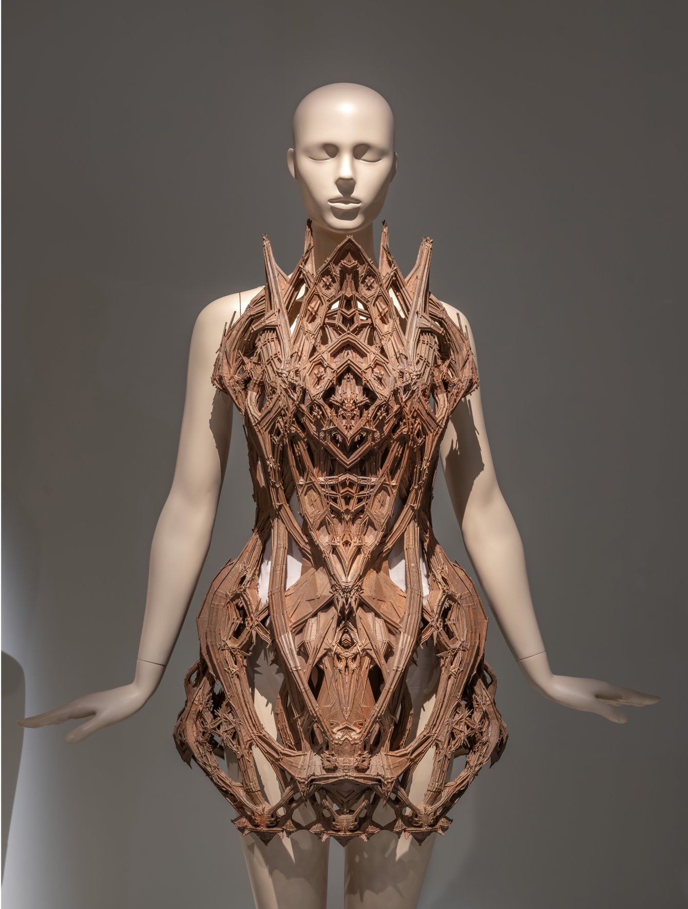 A mannequin wearing a highly structured Iris van Herpen bodice with sharp geometric wooden or copper-toned architectural forms extending across the chest, waist, and hips.
