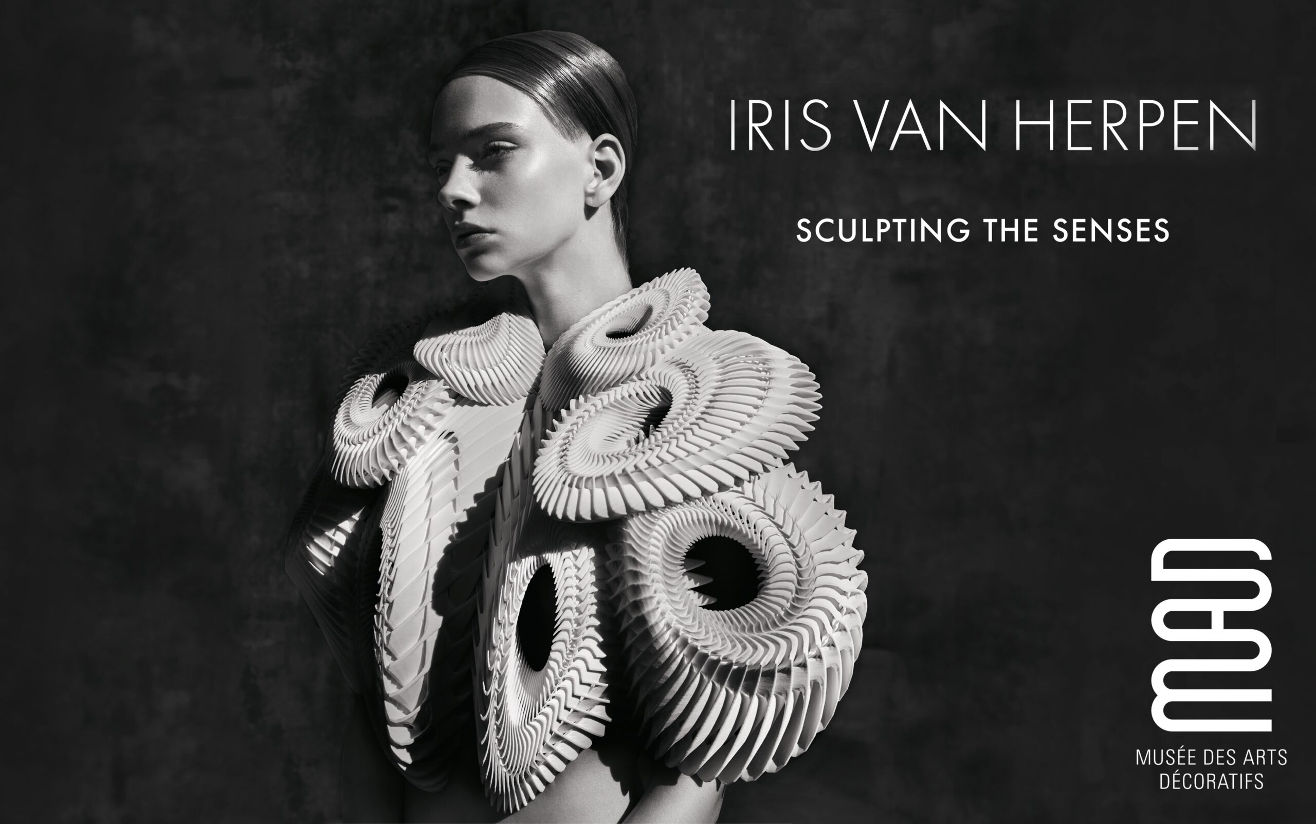 Black-and-white promotional image for Iris van Herpen: Sculpting the Senses showing a model in a sculptural white garment with circular layered forms and the exhibition title text.